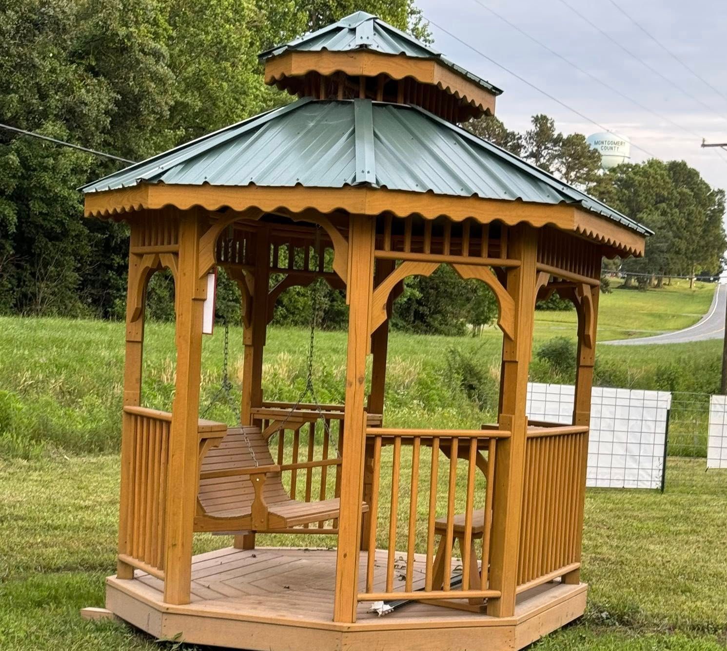 Wooden gazebo with a green roof, and a swing inside, sits on a grassy lawn, and is outside.