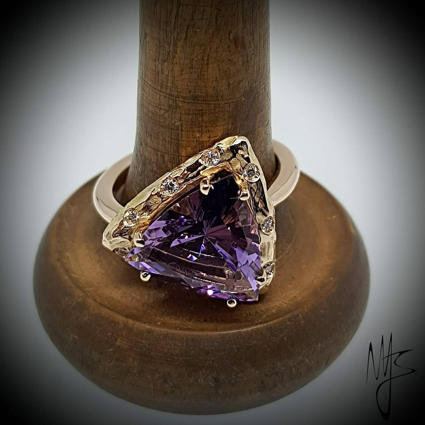 Anello in oro con pietra preziosa triangolare viola, incastonato su un espositore in legno.