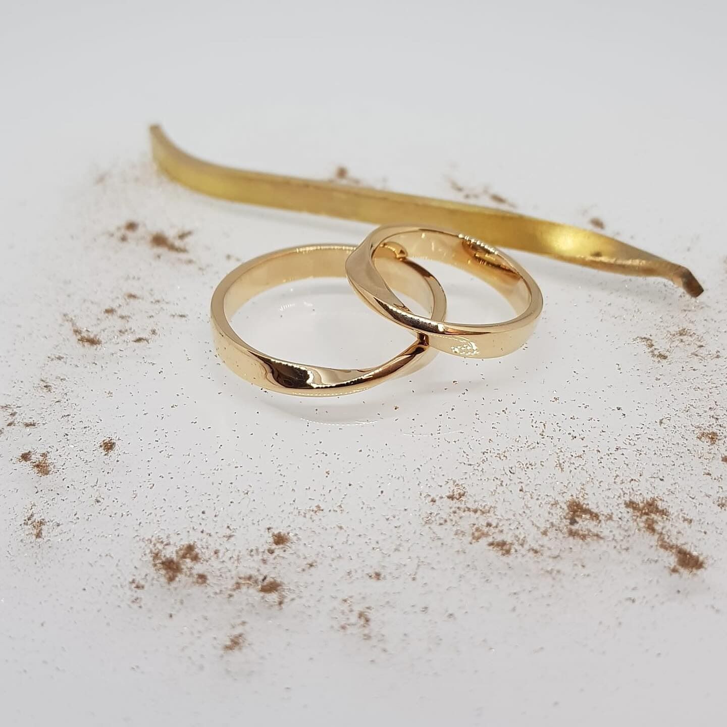 Due fedi nuziali d'oro intrecciate, con un lungo oggetto d'oro ricurvo e particelle marroni sparse.