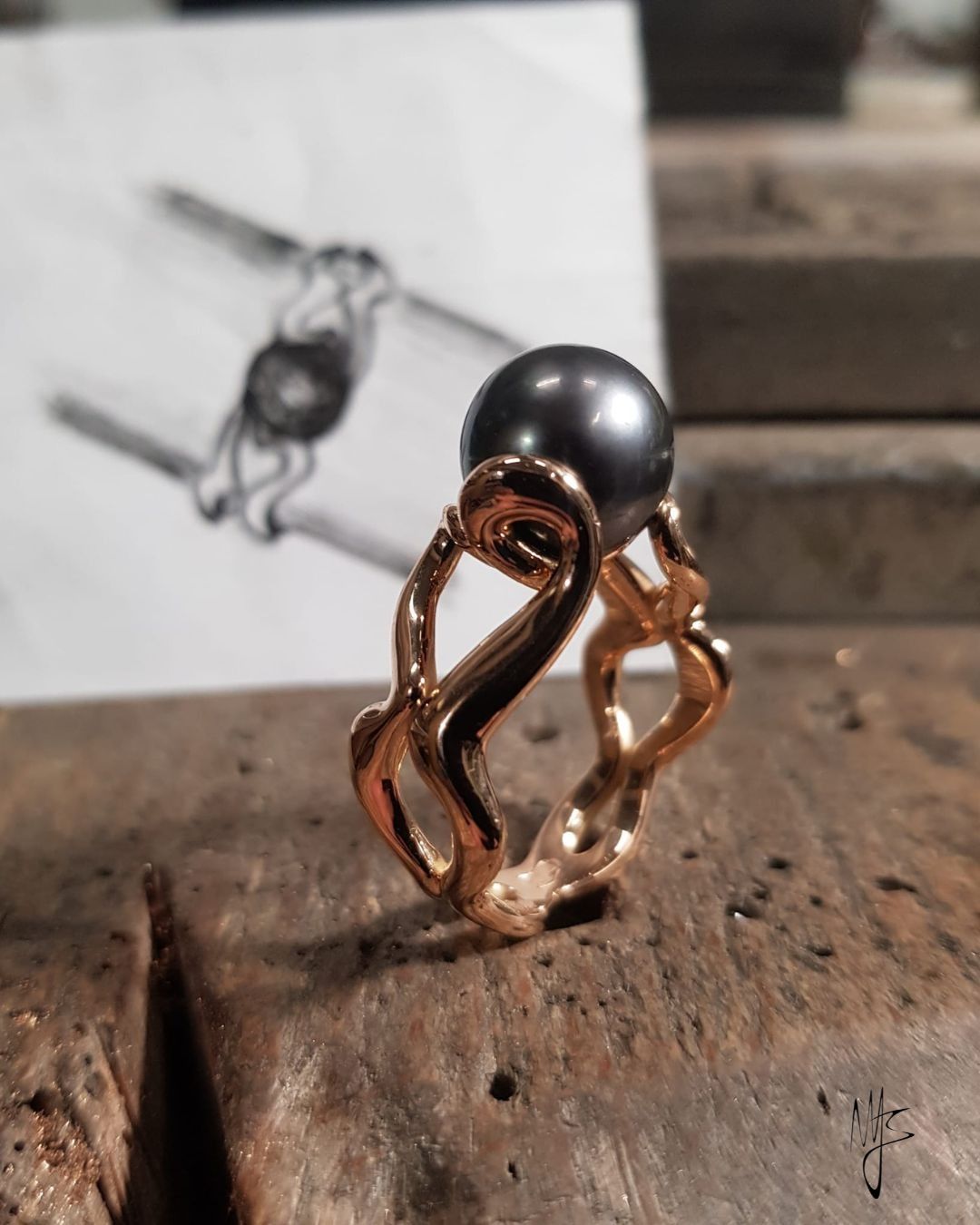Anello in oro con perla scura, accanto a uno schizzo su una superficie di legno.
