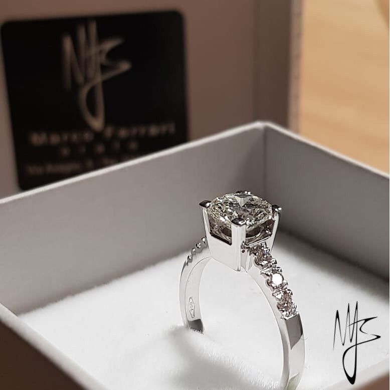 Anello di fidanzamento con diamante in una scatola bianca, che presenta una pietra dal taglio quadrato e una fascia tempestata di diamanti.