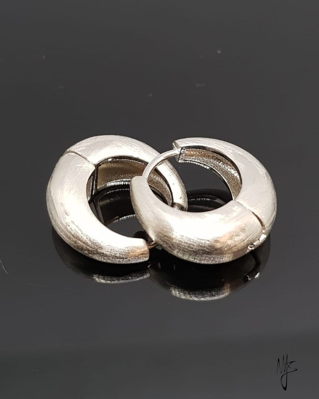 Orecchini a cerchio in argento, finitura opaca, su superficie nera riflettente.