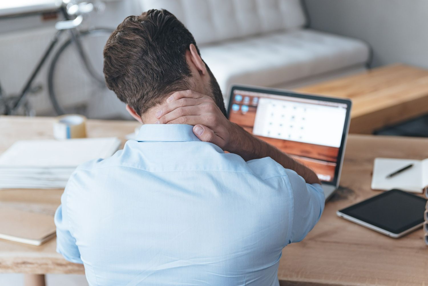 A man is sitting at a desk with a laptop and holding his neck.