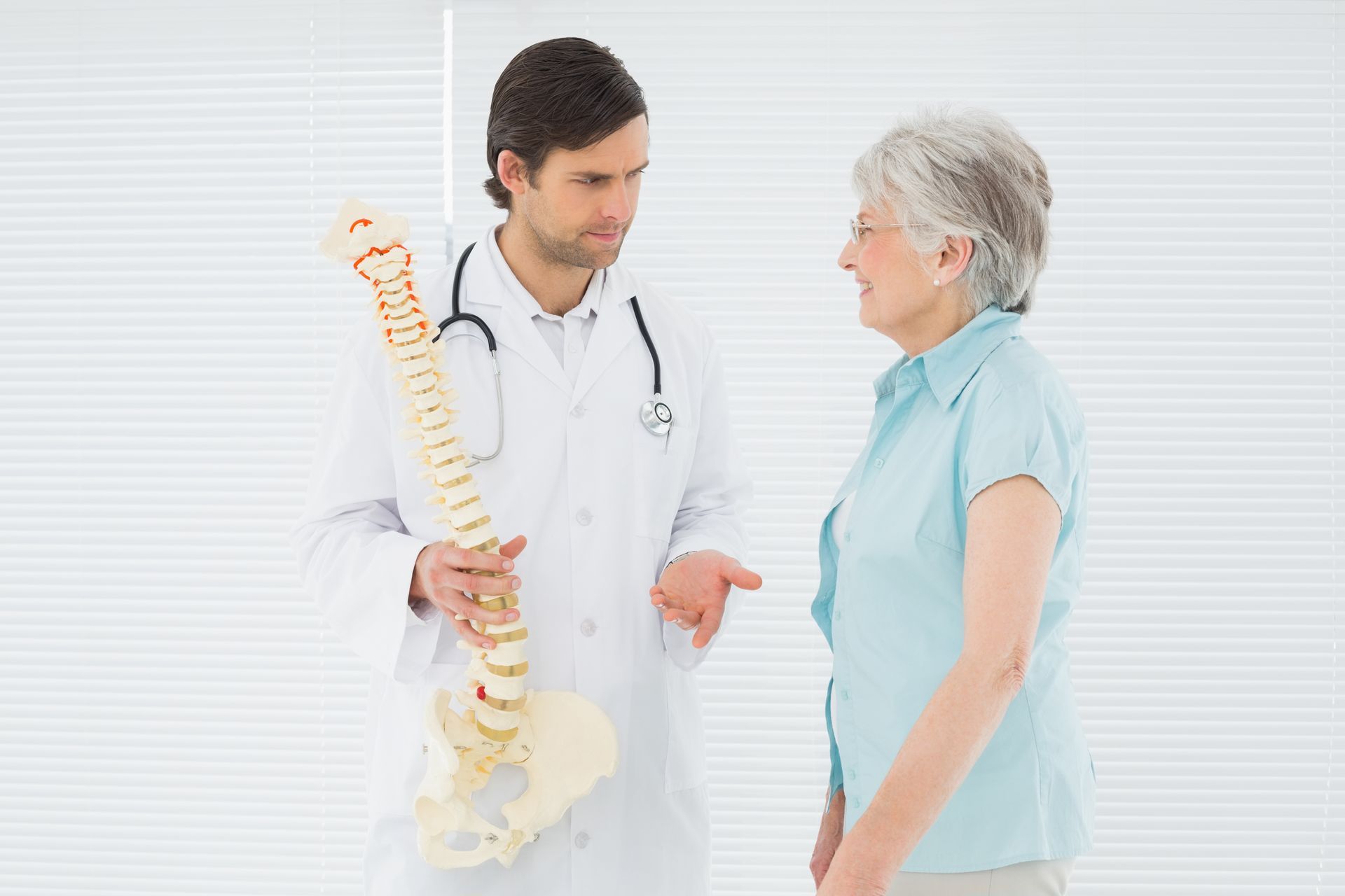 A doctor is talking to an elderly woman while holding a model of a spine.