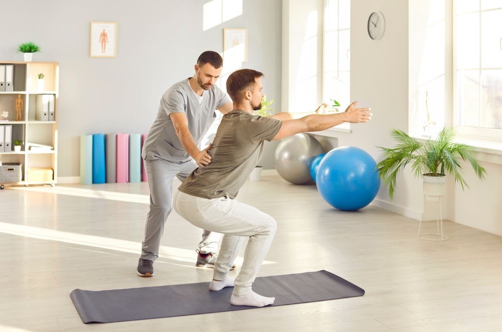 A man is squatting on a yoga mat while a doctor helps him.