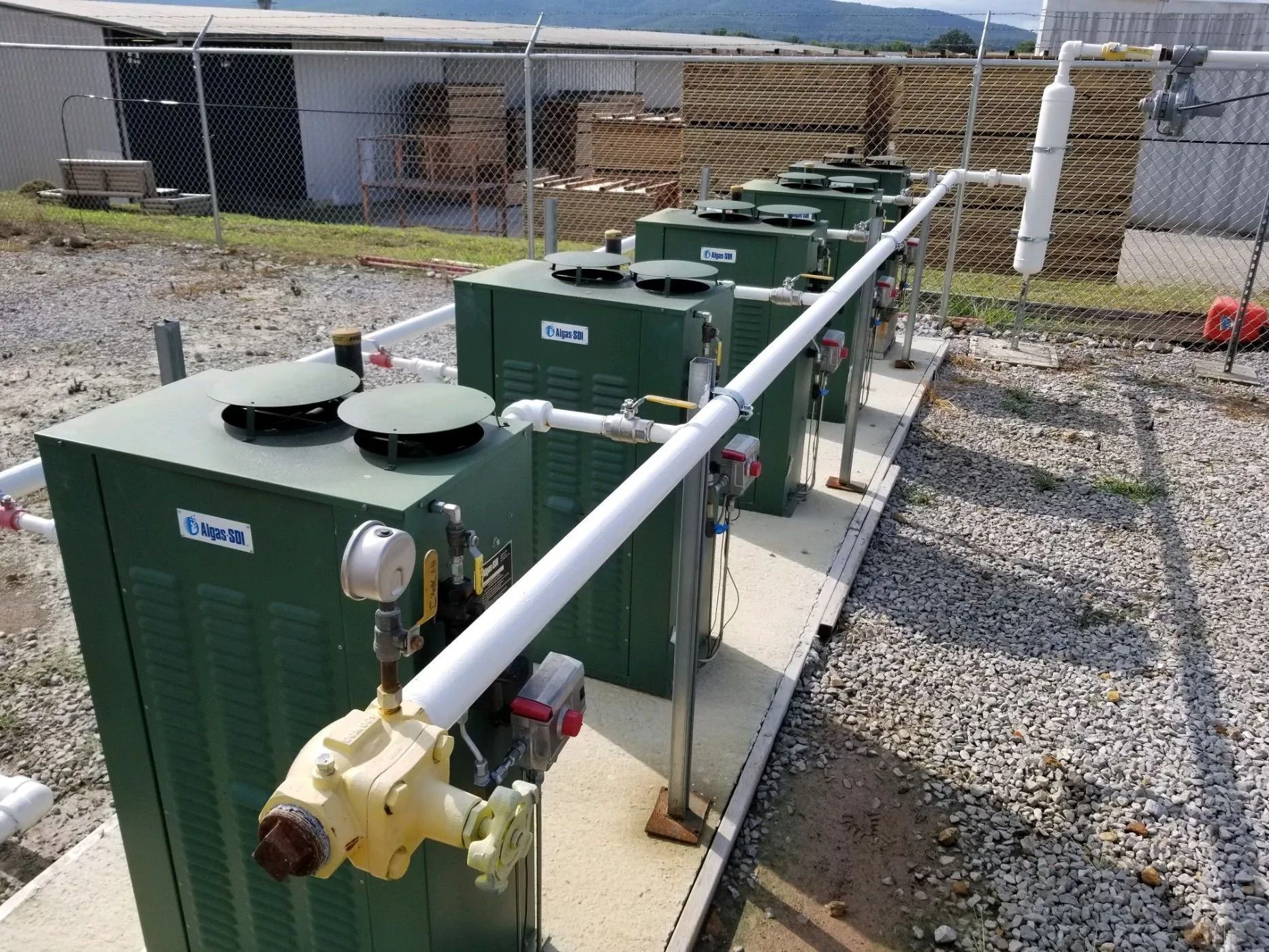 Row of green industrial units with piping outdoors on a concrete pad, surrounded by a chain-link fence.