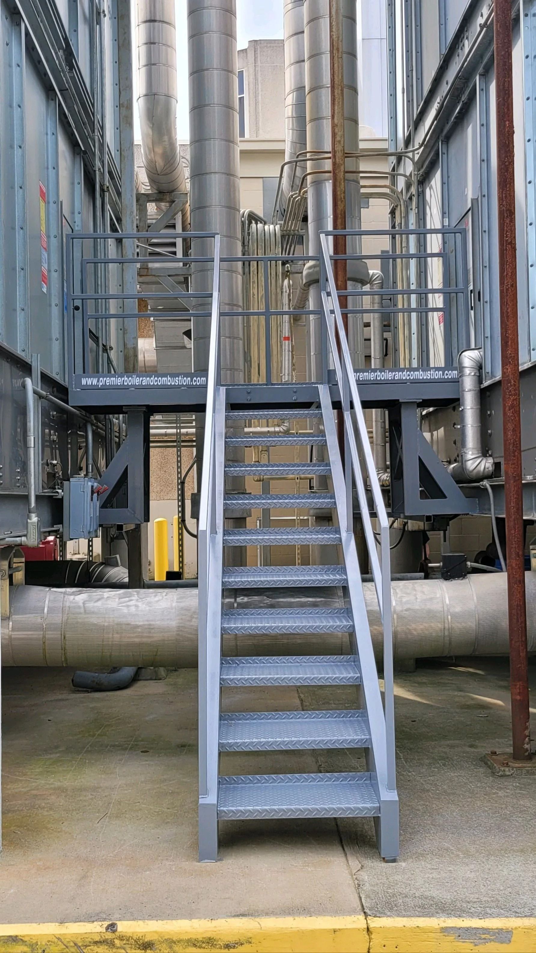 Metal staircase leading up to a platform between industrial buildings, with pipes and equipment visible in the background.
