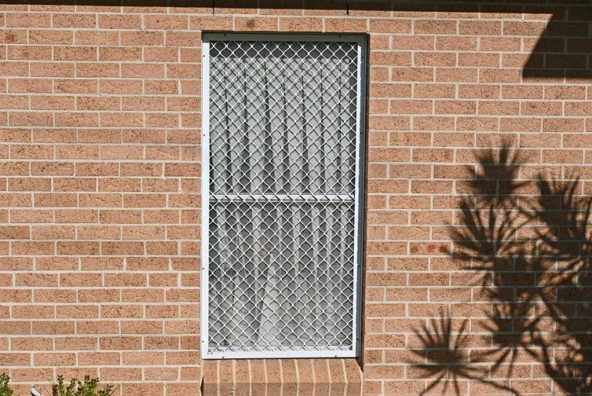 A Brick Wall With a Window With a Chain Link Grille Fence Around It — Affordable Blinds, Doors & Security Pty Ltd in Tweed Heads South, NSW