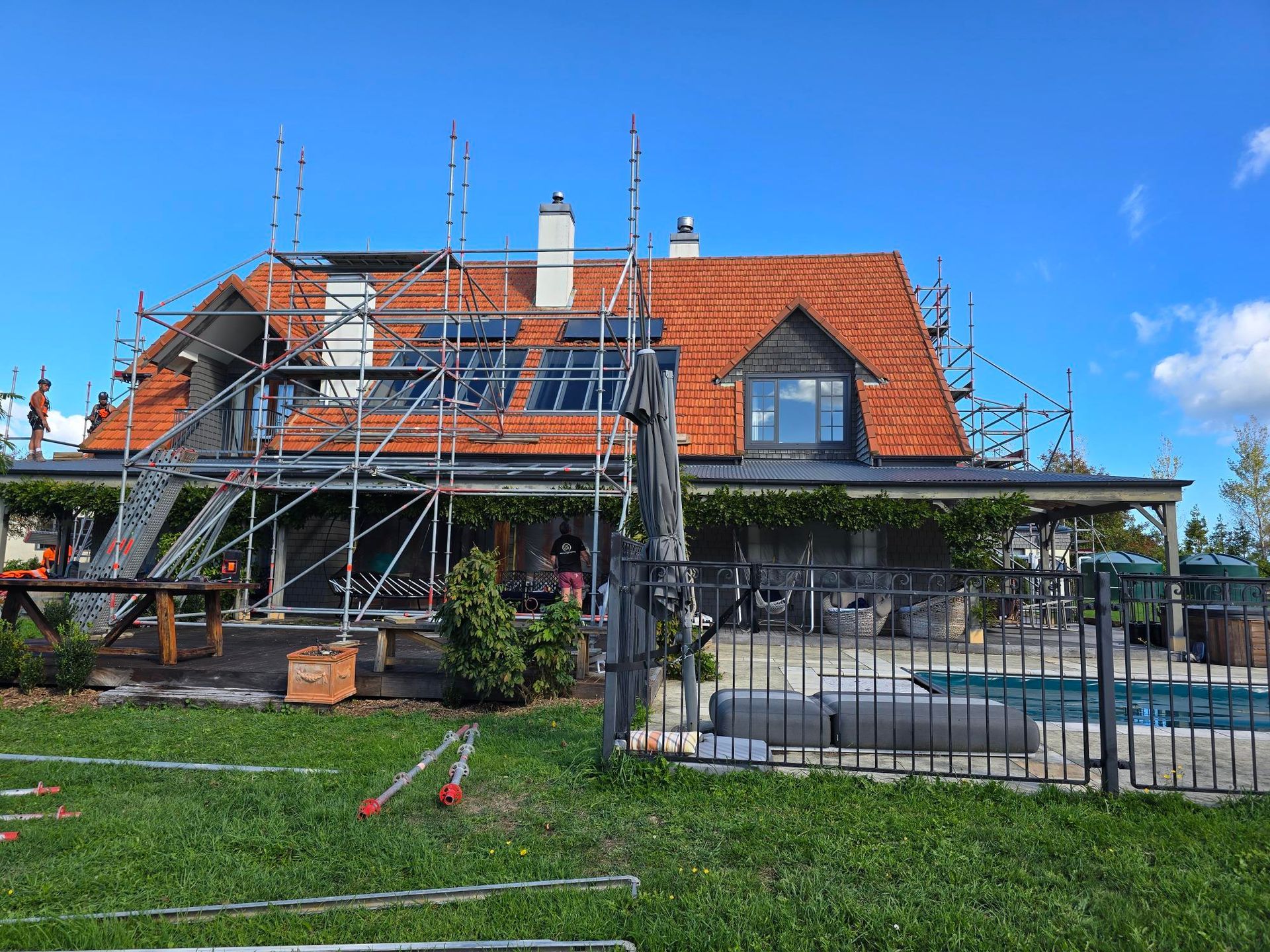 House with red tiled roof, scaffolding, and solar panels under installation. Green grass and blue sky.