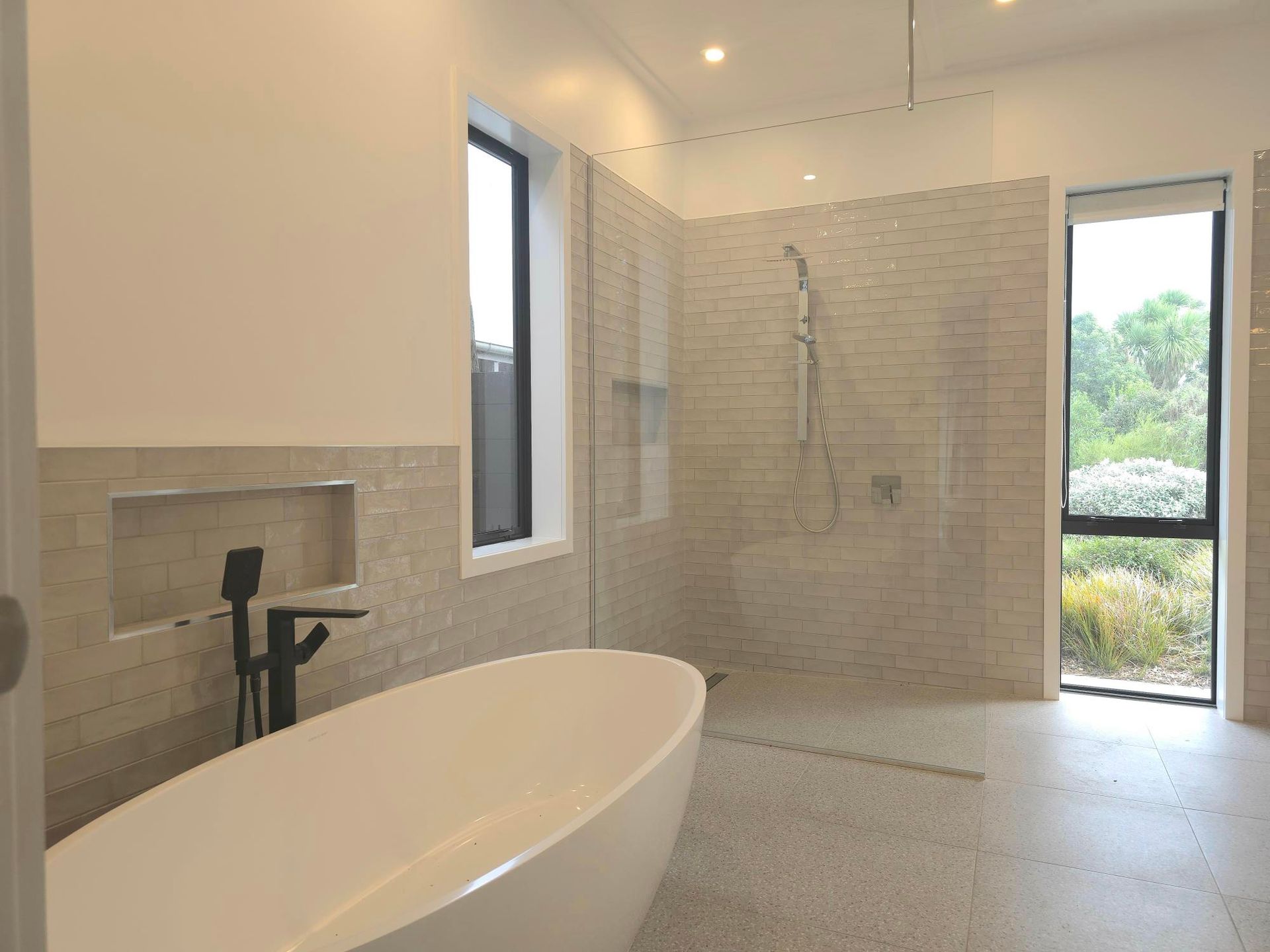 Modern bathroom with a freestanding tub, shower, and window. Light-colored walls and floors.
