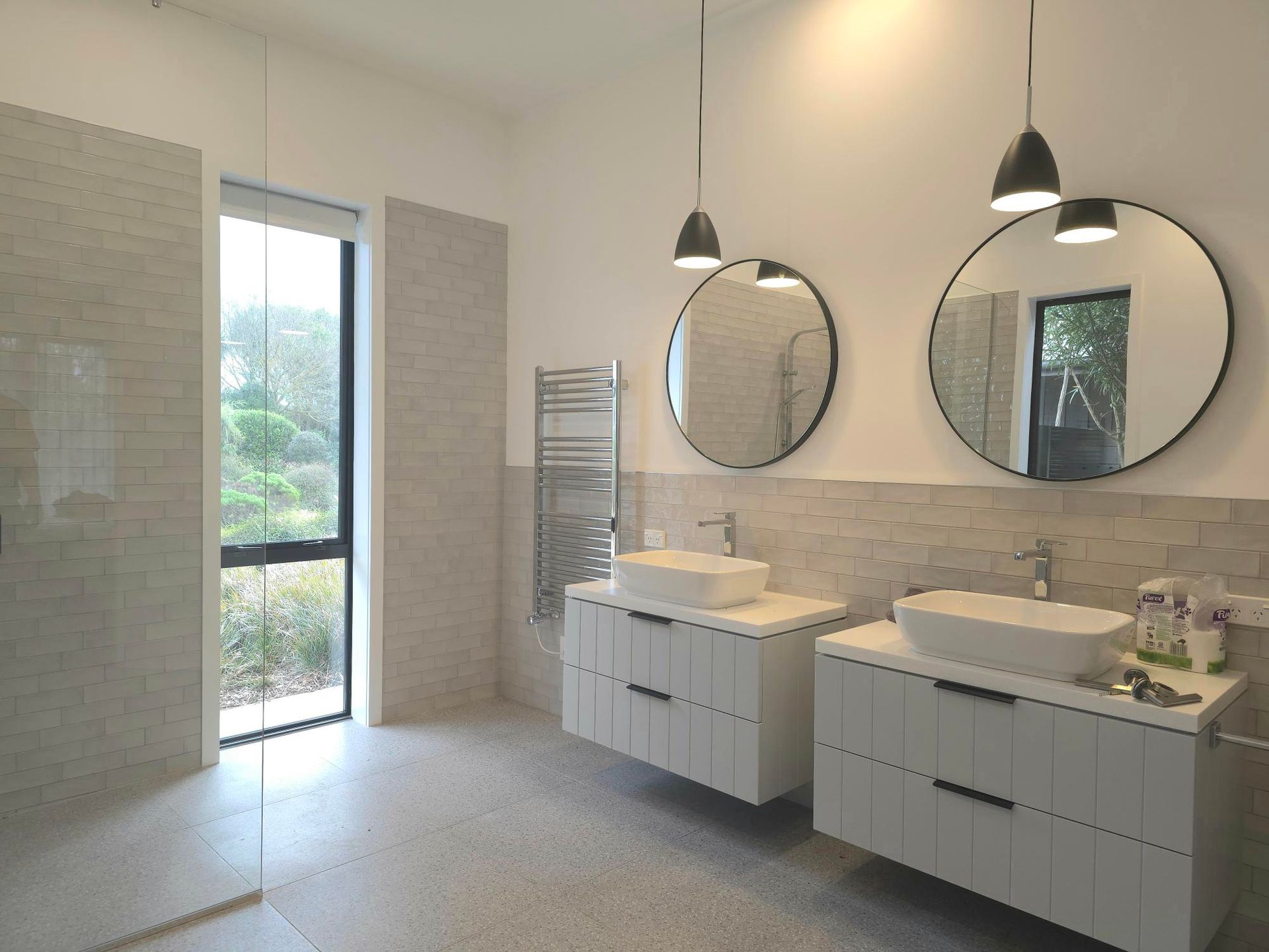 Modern bathroom with two white vanities, round mirrors, and a large window overlooking nature.