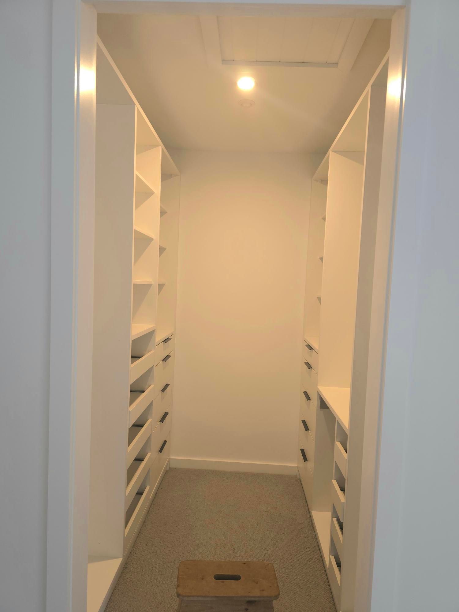 Narrow, white walk-in closet with shelves and drawers on both sides. A small stool sits on the gray carpet floor.