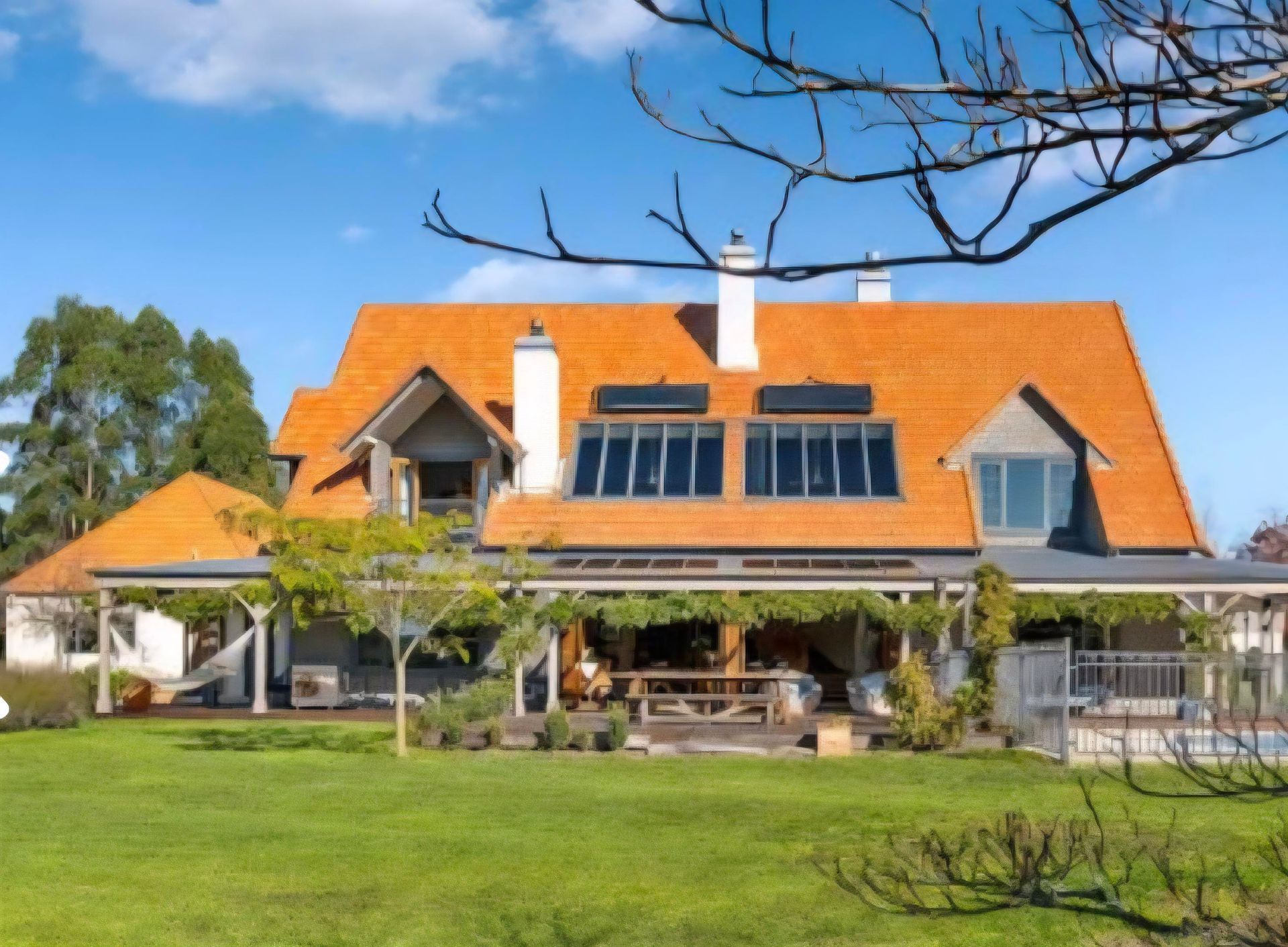 Large house with orange roof, solar panels, and a wraparound porch on a sunny, green landscape.