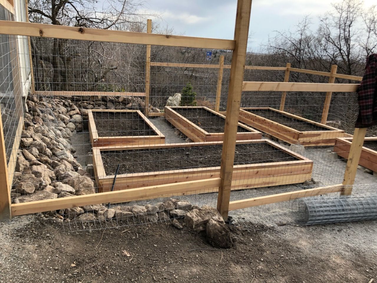 Raised wooden garden beds under construction beside a rock wall and fence in a backyard.