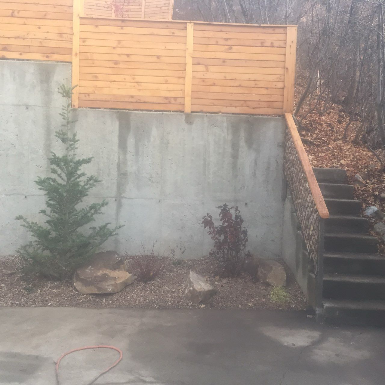 Concrete retaining wall with wooden fence, small shrubs, and stairs beside a driveway.