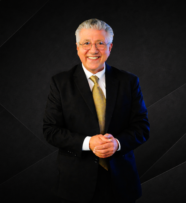 Man in a black suit with a gold tie, smiling with hands clasped, against a dark backdrop.