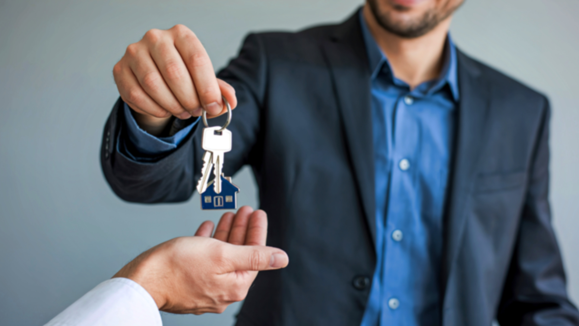 Man in suit handing keys with house charm to another person.