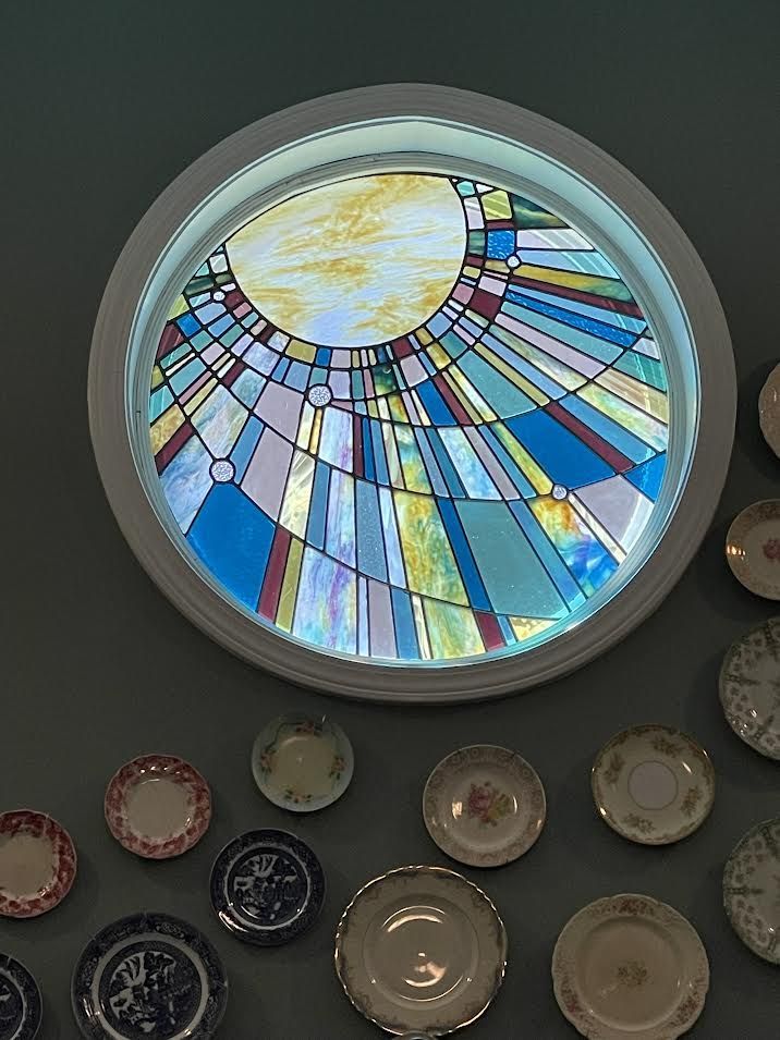 Round stained-glass window with sunburst design above a collection of decorative plates on a grey wall.