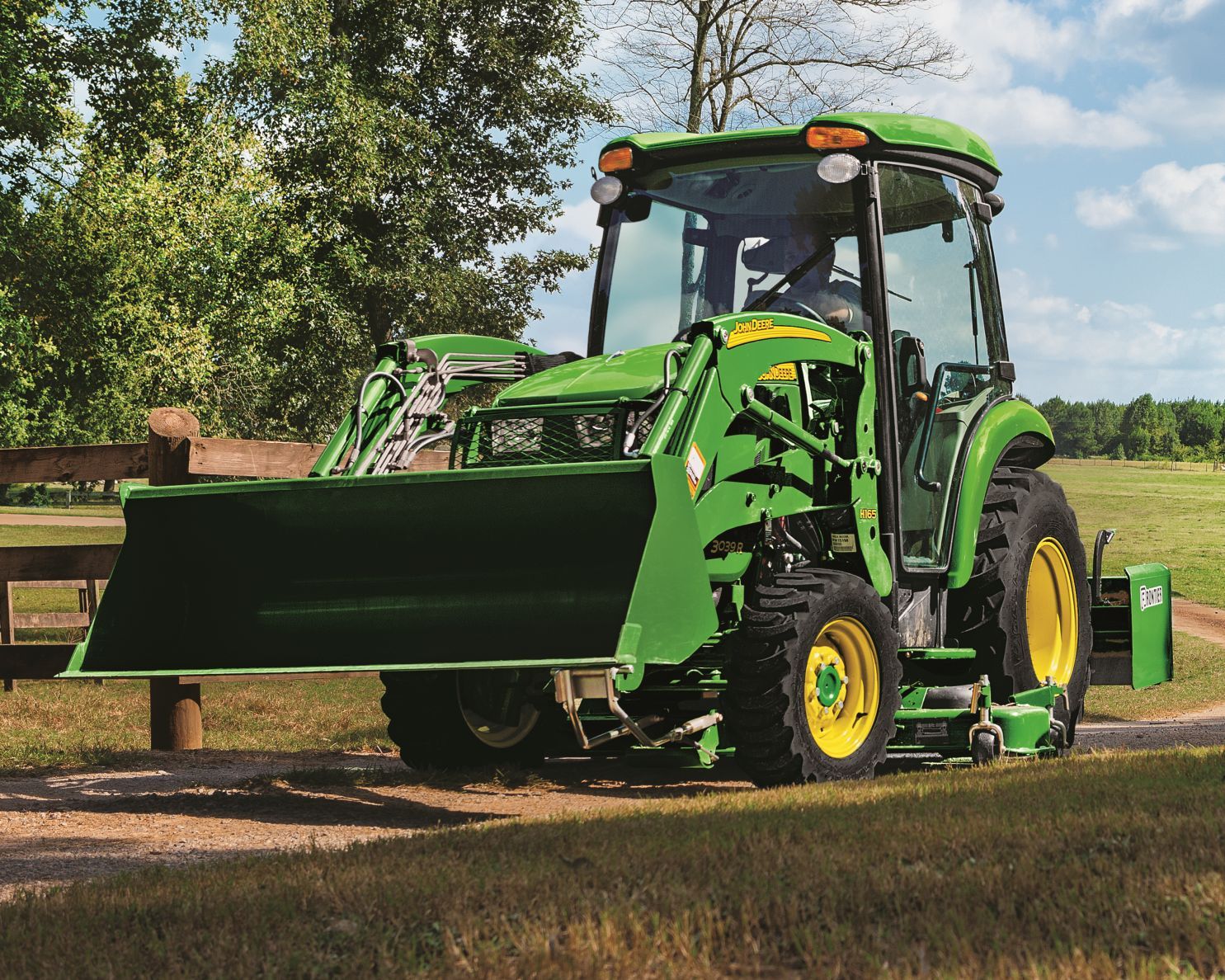A person in blue overalls and protective eyewear repairs the engine of a green tractor in a field. A person in blue overalls and protective eyewear repairs the engine of a green tractor in a field.