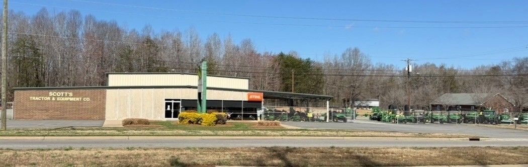 A wide-angle shot of a brick commercial building with a tan facade, green signage, and a yard filled with farm equipment.