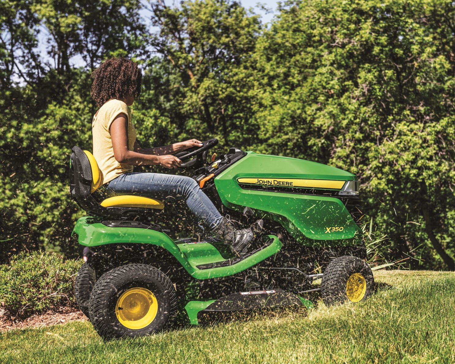 A person in blue coveralls uses a screwdriver to work on the engine of a green tractor inside a repair shop.