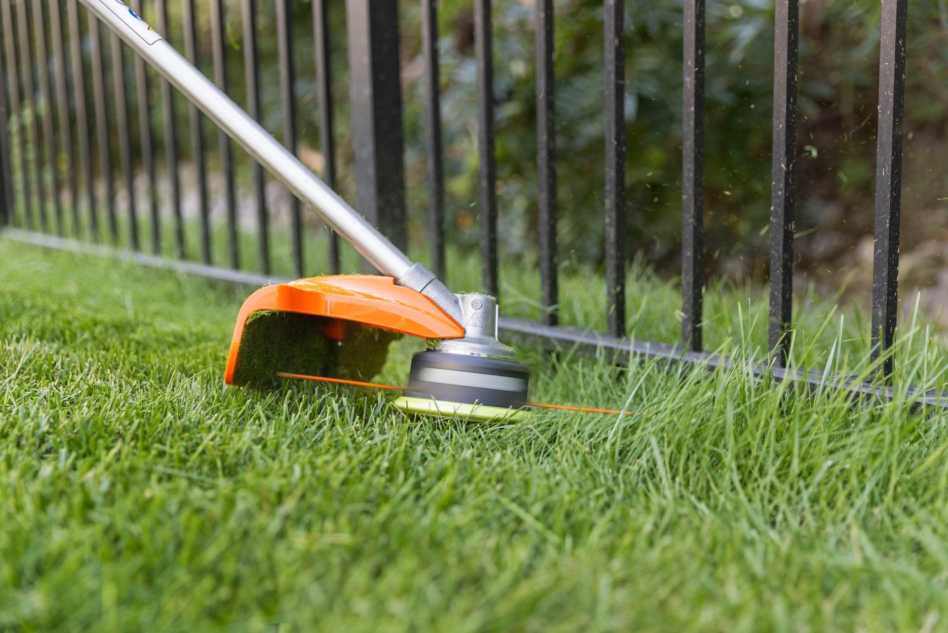 A person uses an orange handheld weed trimmer to cut grass along a wooden fence.