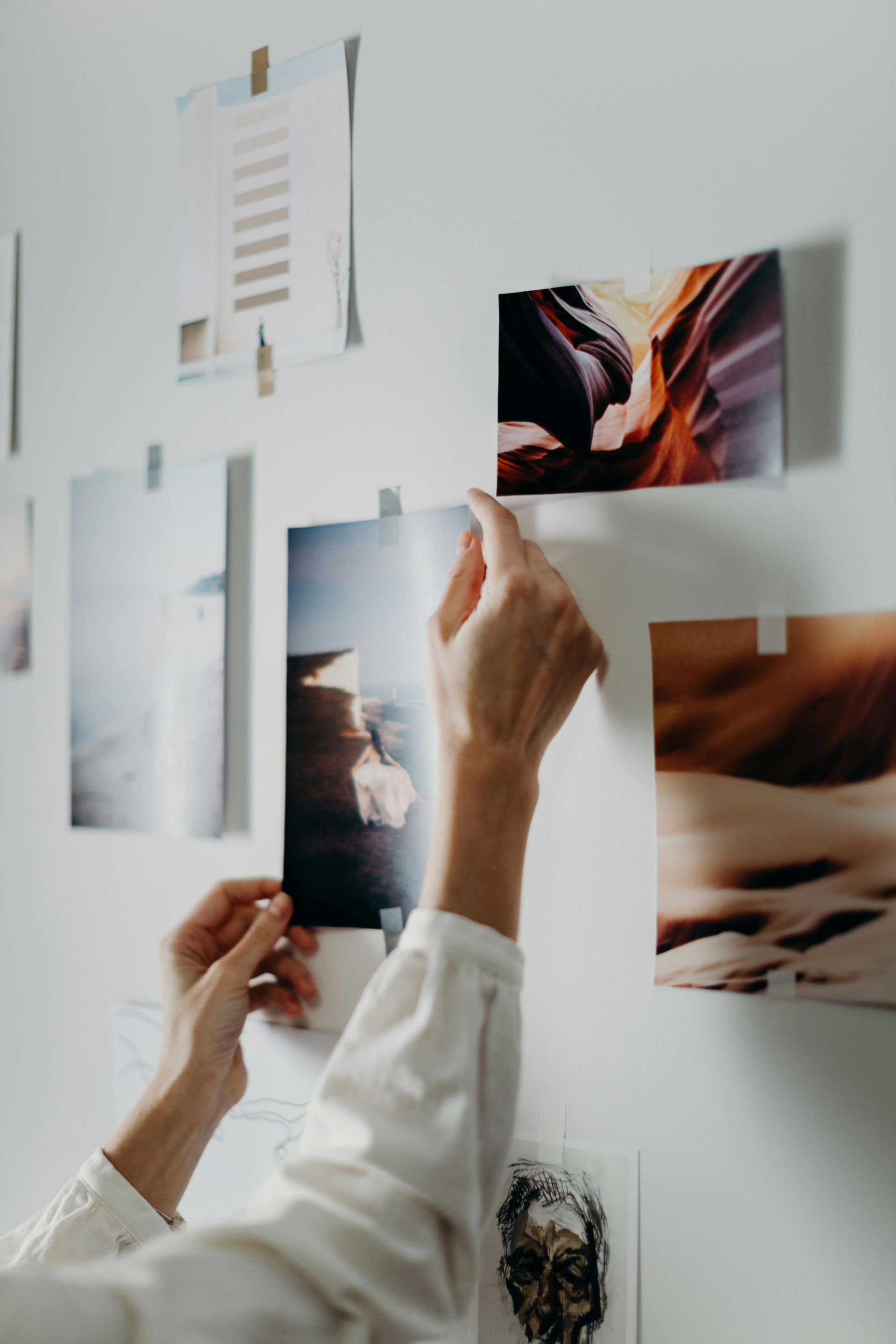 Des mains fixent des photos sur un mur blanc avec du ruban adhésif. Les images représentent des paysages aux tons chauds.