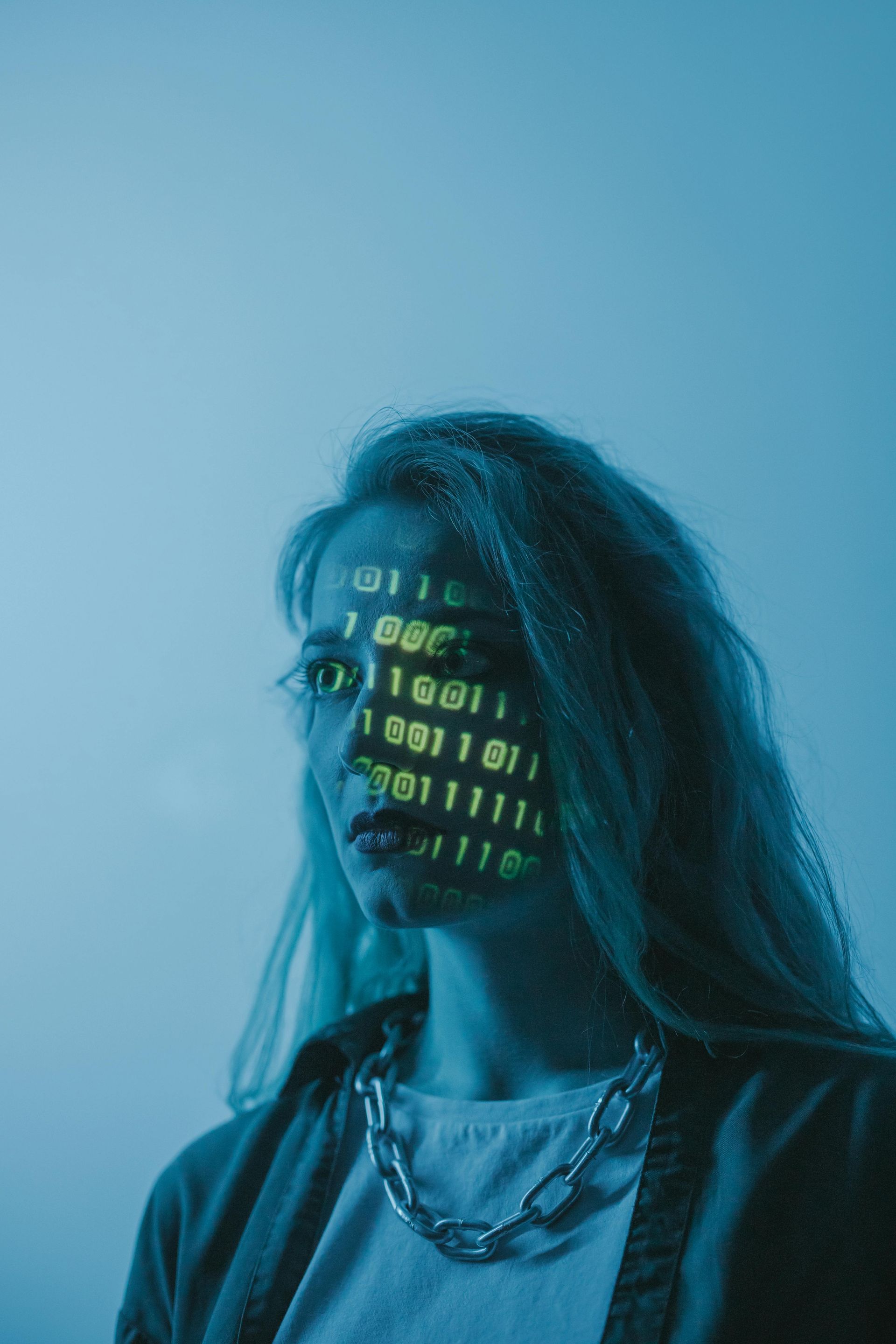 Visage de femme avec un code binaire projeté dessus, portant une chaîne, éclairé par une lumière bleue.