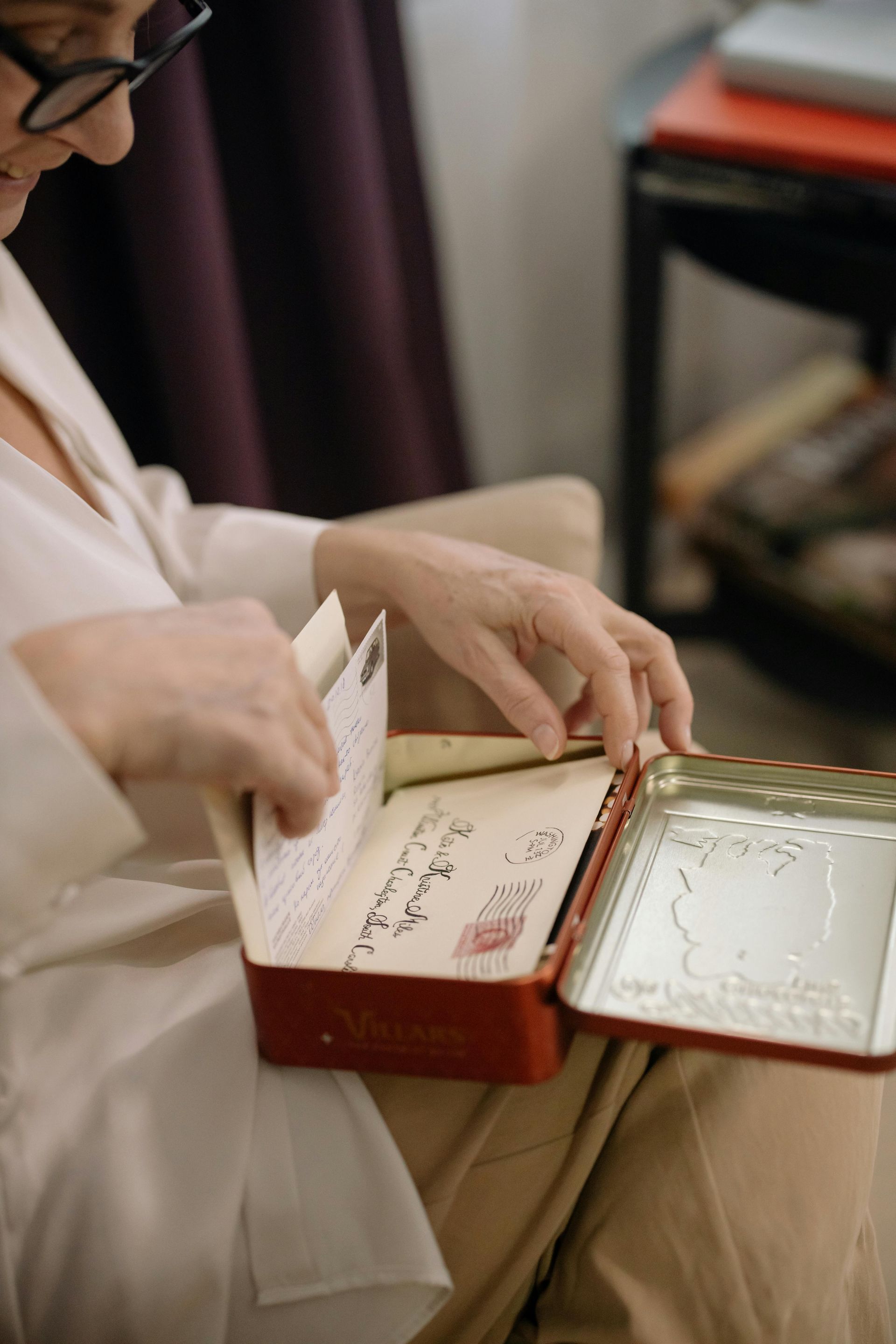 Tiiming au coeur de votre histoire familiale Femme ouvrant une boîte rouge et examinant de vieilles lettres. Assise à l'intérieur, portant des lunettes et une chemise blanche.