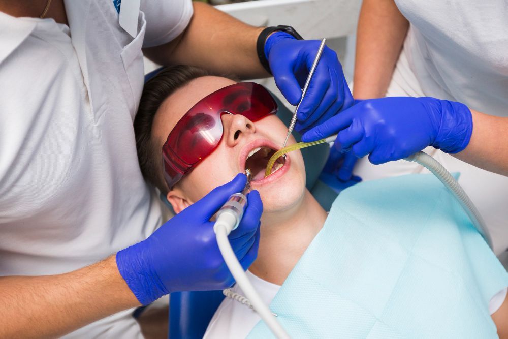 Person with mouth open at dentist; assistants with tools, wearing blue gloves and protective glasses.