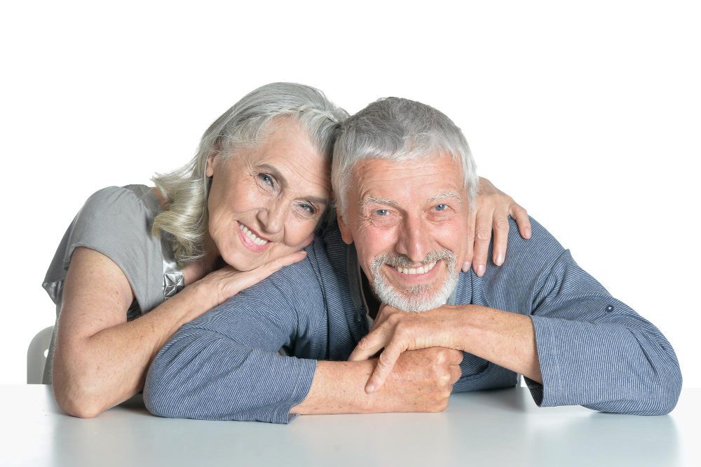 Smiling senior couple leaning on a white surface. Woman's arm around man. Both have gray hair.