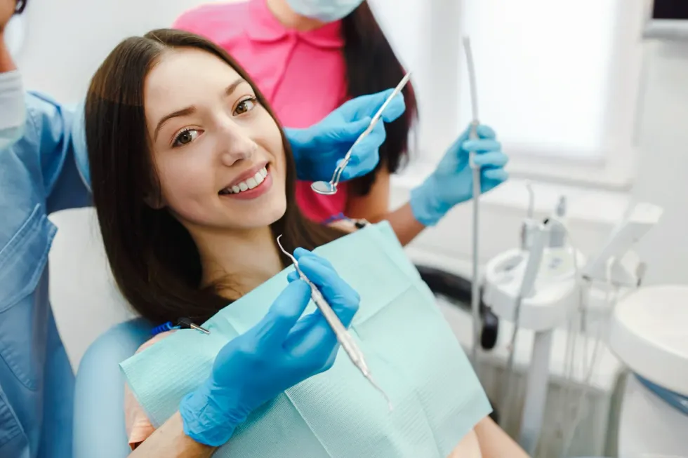 Woman smiles in a dentist's chair while hygienists hold tools. Bright setting, blue gloves.