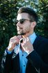 Man in sunglasses adjusts his shirt collar, wearing a blue suit jacket, in front of green foliage.