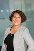 Smiling woman in business attire, short curly hair, standing near glass wall.