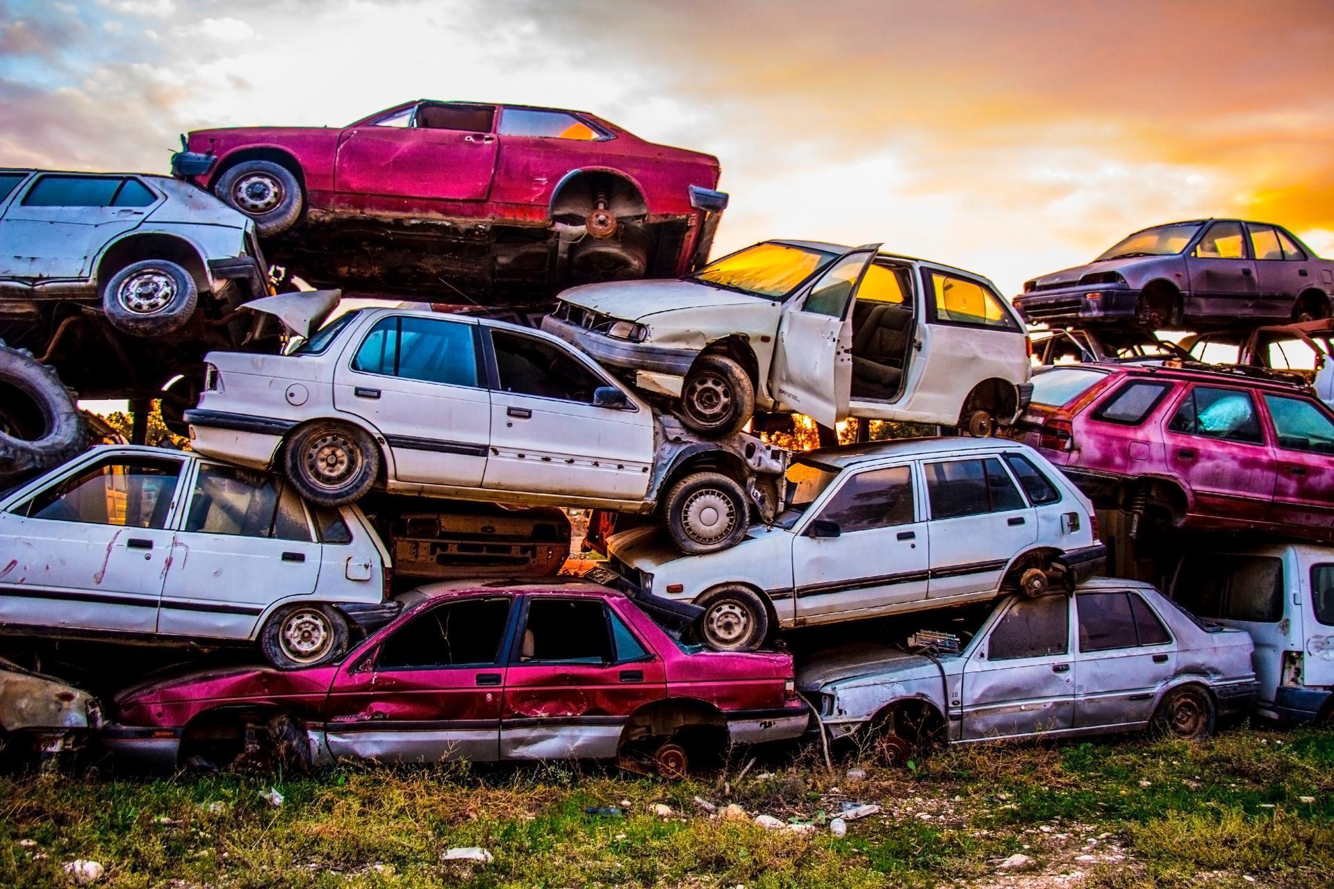 Stack of crushed and junk cars piled in scrapyard under colorful sunset sky.