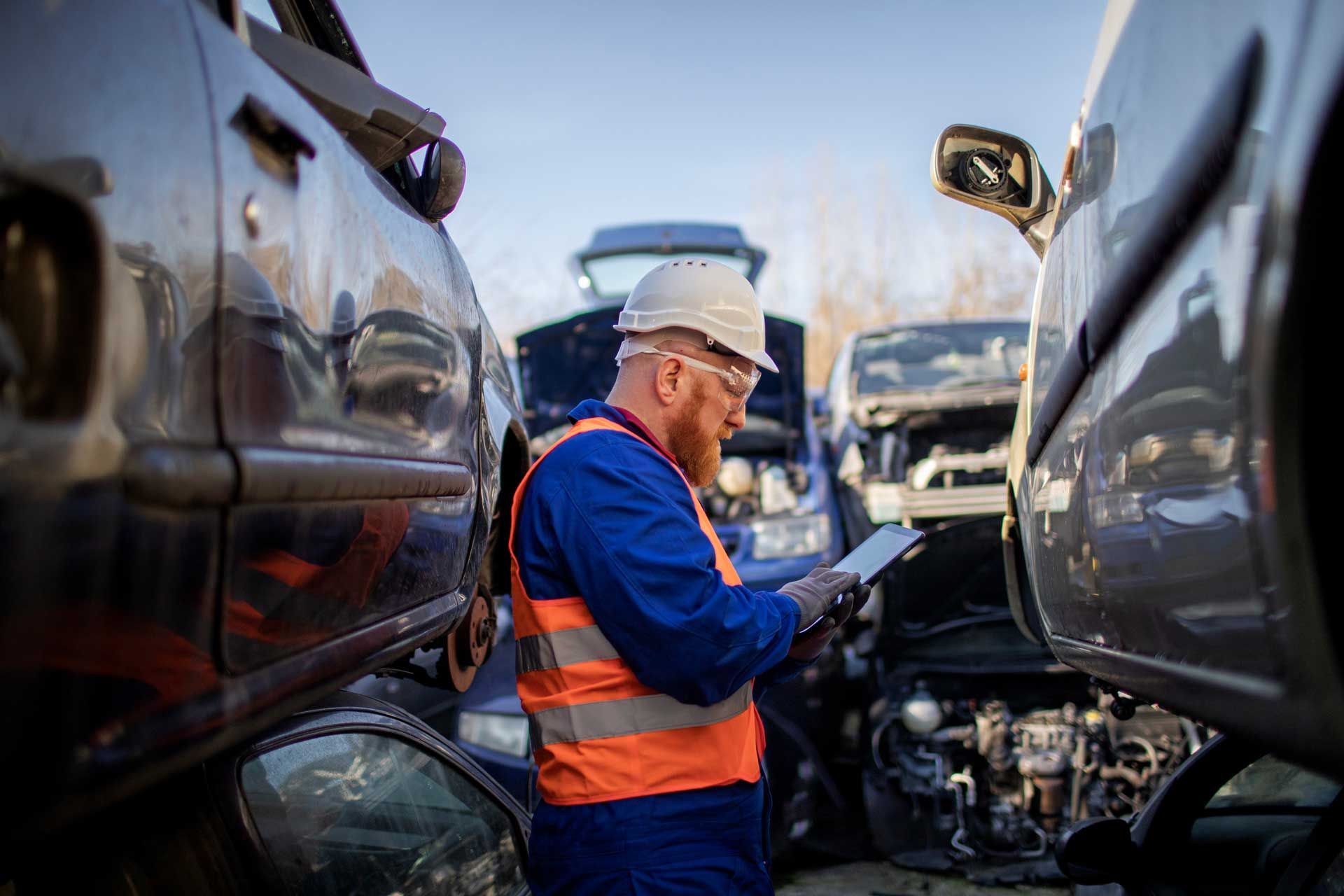A mechanic in a junkyard uses a tablet to inspect a car engine, wearing safety gear.