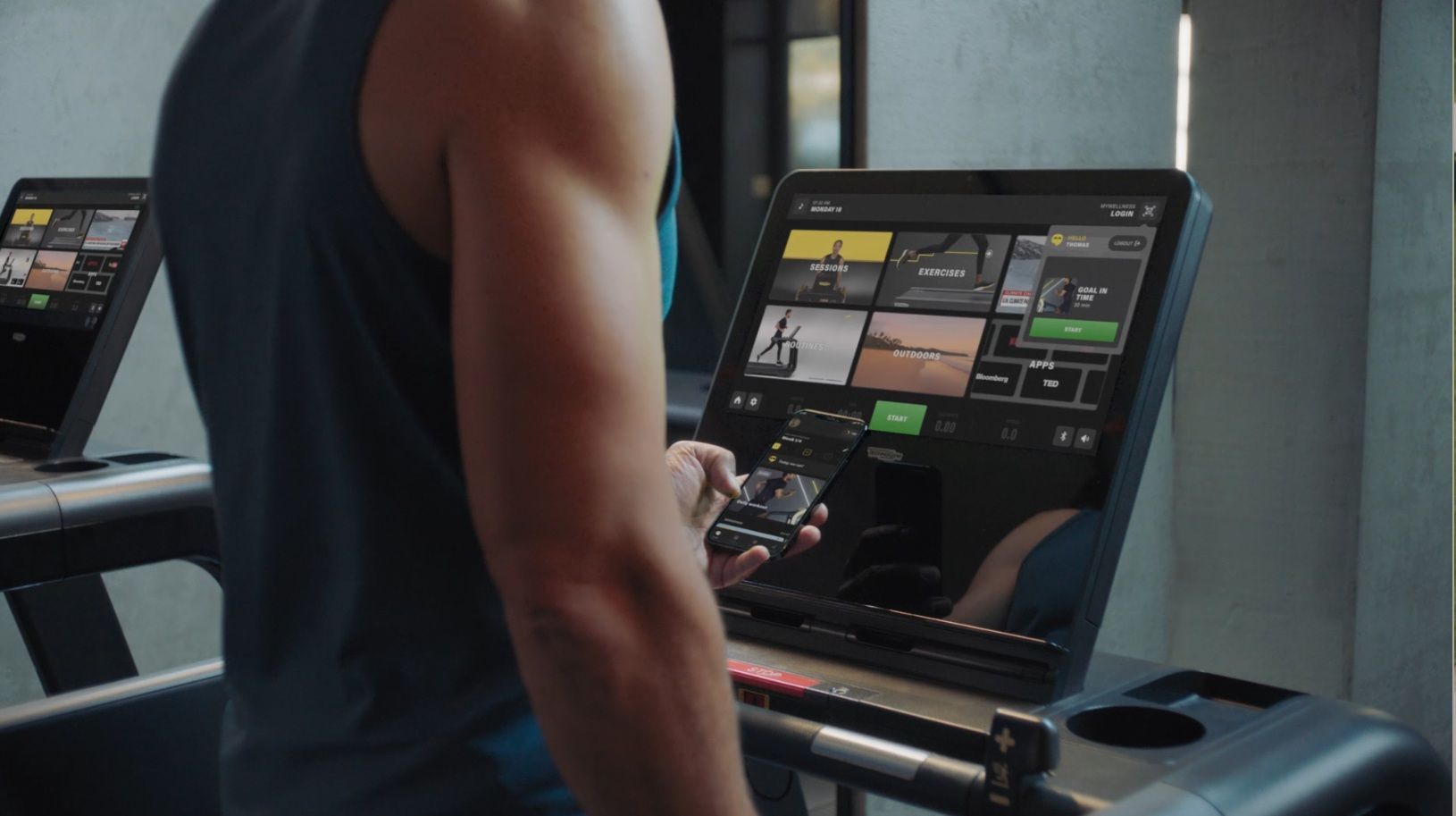 A person in a gym interacts with a smartphone while standing before a treadmill featuring a touchscreen interface.