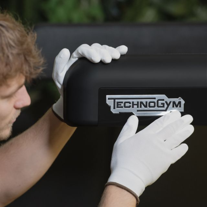 Hands in white gloves carefully touch the metallic Technogym logo on a black fitness equipment surface.