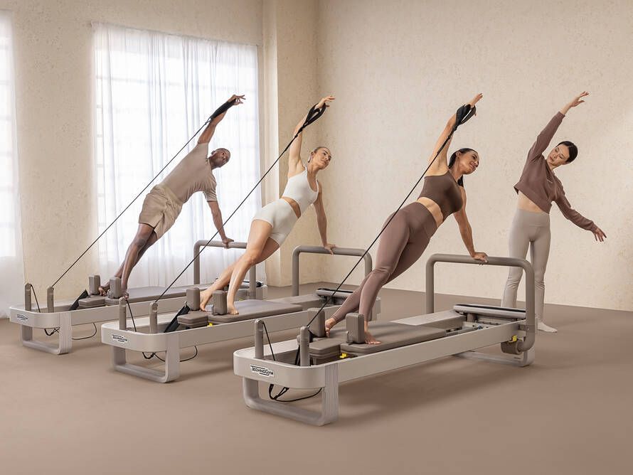 Four people in neutral activewear performing a side plank exercise on Pilates reformers in a bright, minimalist studio.