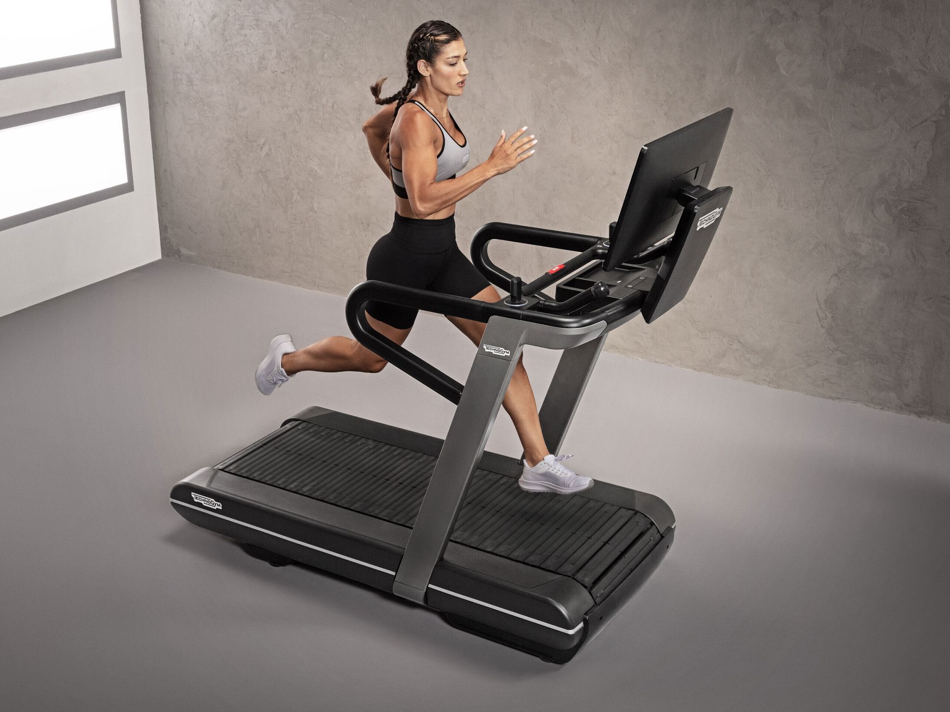 A person running on a treadmill with a digital display screen, set against a plain grey studio backdrop.