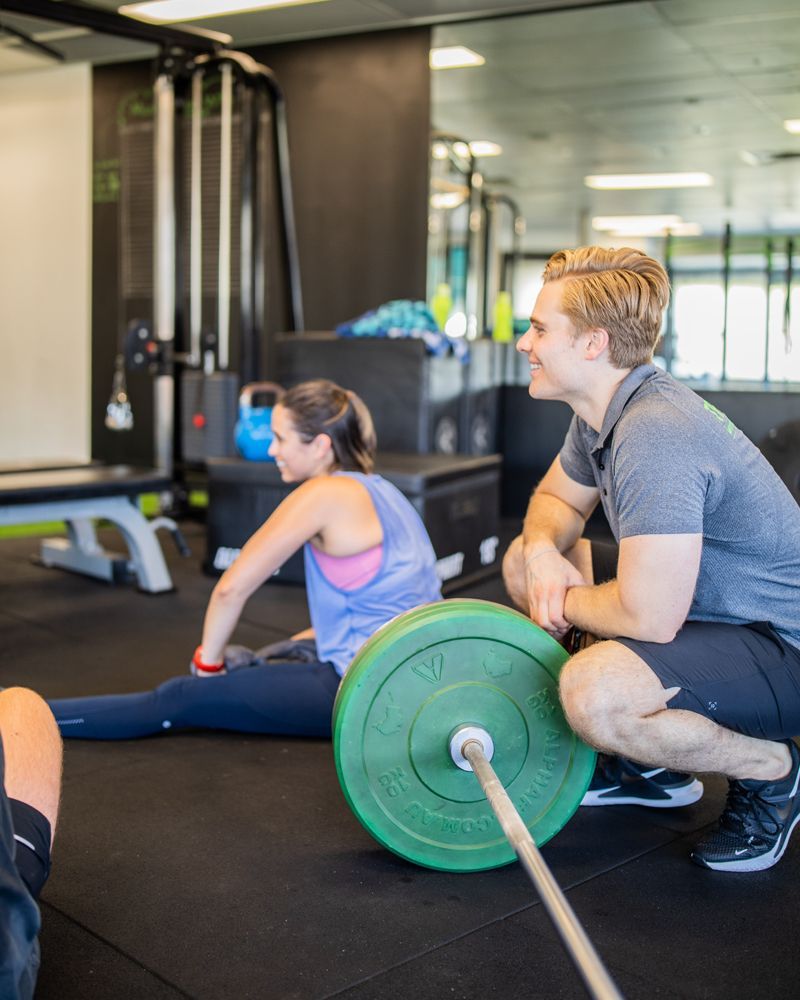 A man is kneeling down next to a green barbell in a gym.