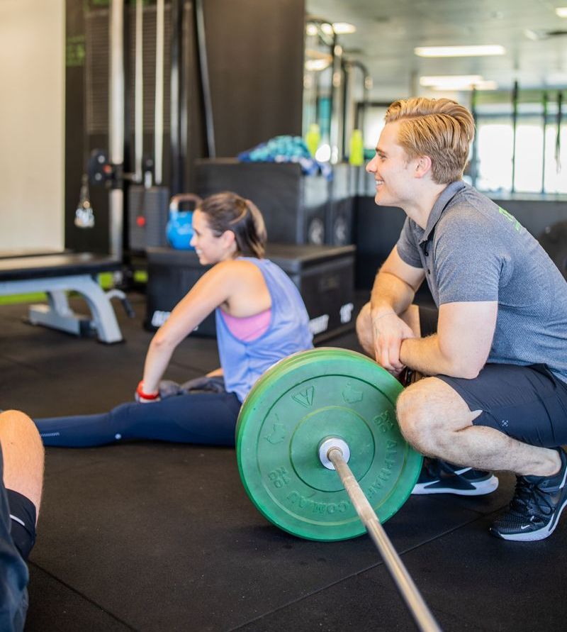 A man is squatting down next to a green barbell in a gym.