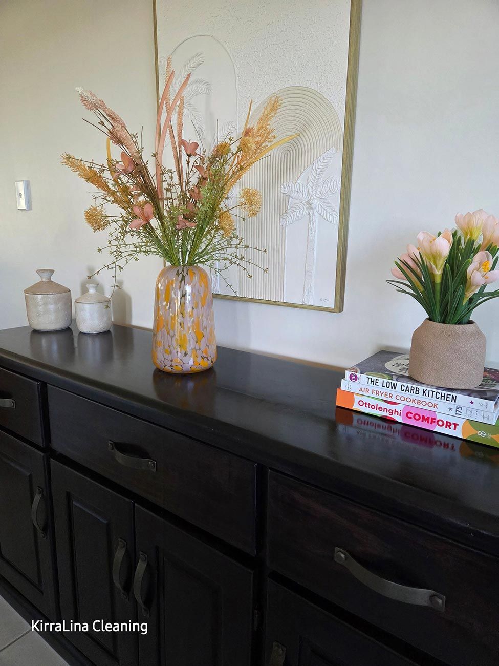 Dark Wood Cabinet with Vases of Flowers, Books, and Decor Against a Wall with Art — KirraLina Cleaning Services in Tweed Heads, NSW