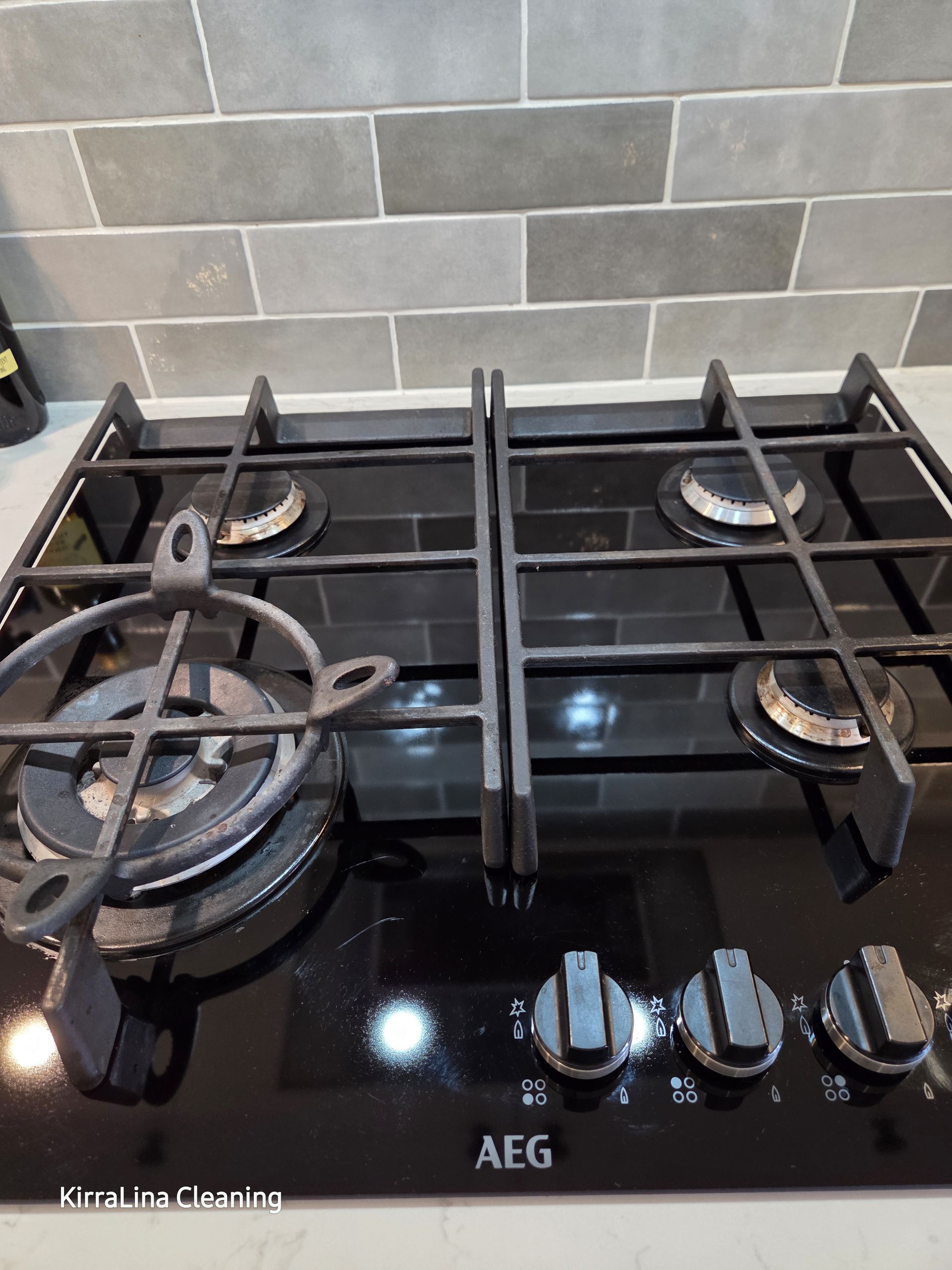 Black glass AEG gas stovetop with four burners and cast-iron grates in a tiled kitchen. — KirraLina Cleaning Services in Tweed Heads, NSW