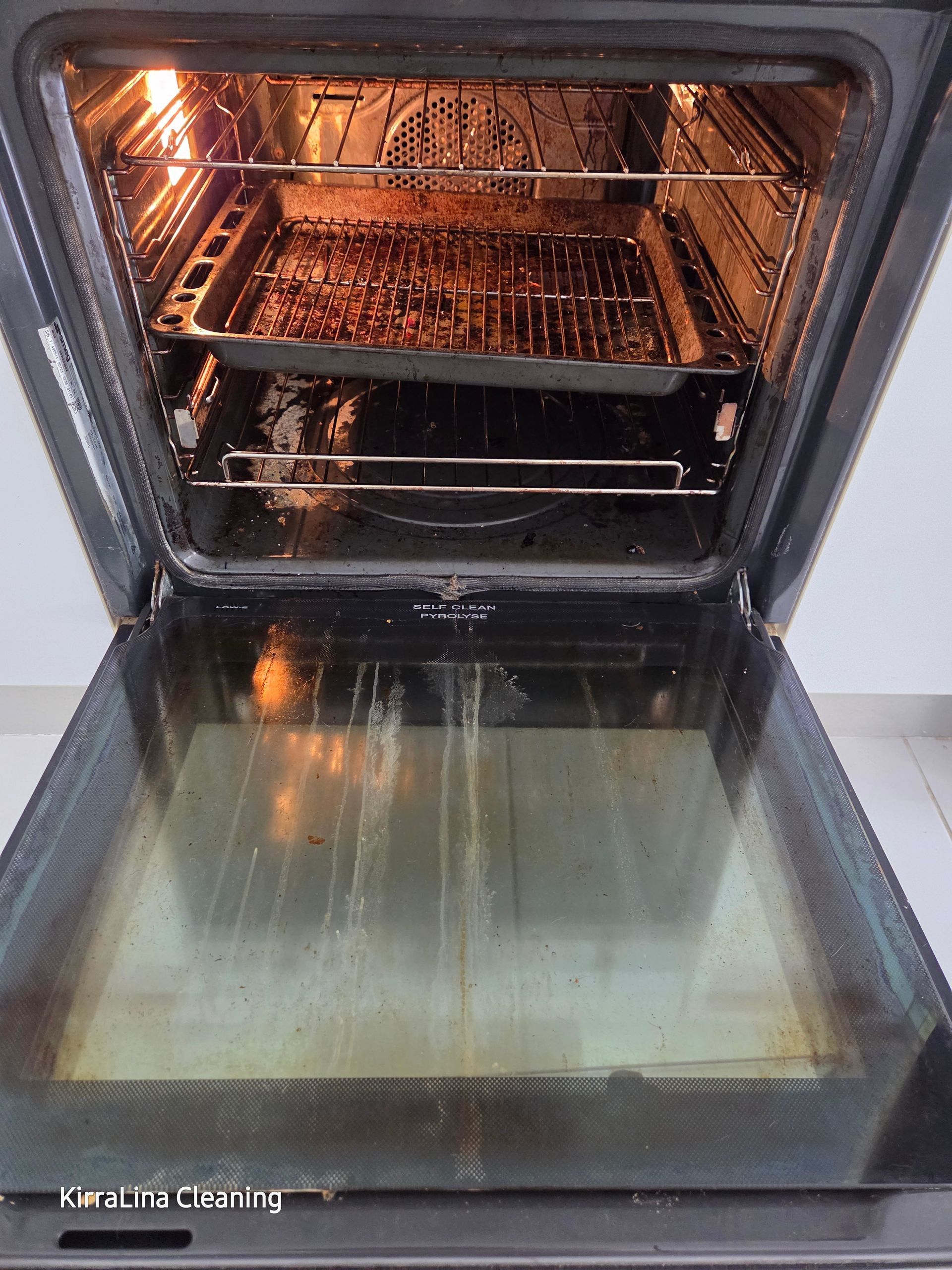 An open oven interior with a dirty baking tray and wire rack inside, and visible streaks on the oven door glass — KirraLina Cleaning Services in Tweed Heads, NSW