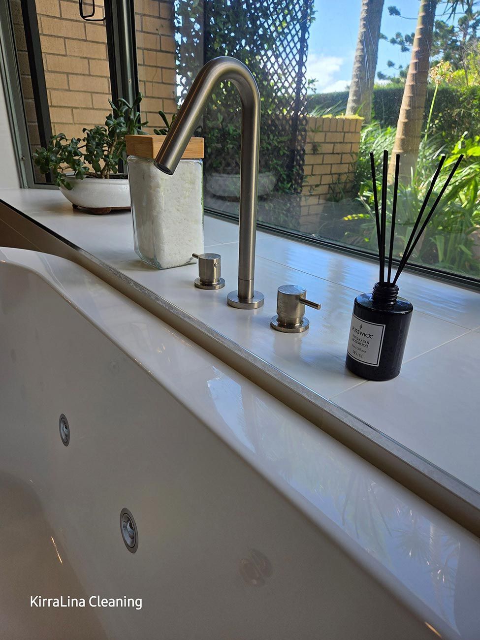 A bathtub faucet with matching handles, next to a window overlooking greenery. A reed diffuser and potted plant sit on the windowsill.