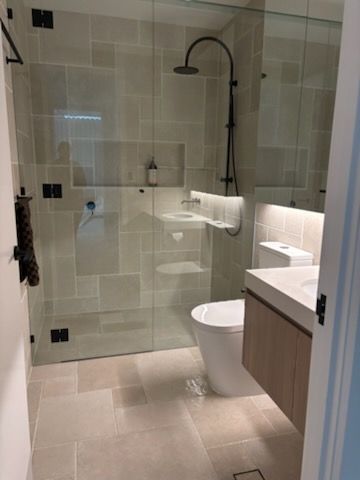 A Modern Bathroom with A Glass Shower, Toilet, and Vanity with A Light-Colored Stone Tile Floor and Walls — KirraLina Cleaning Services in Tweed Heads, NSW