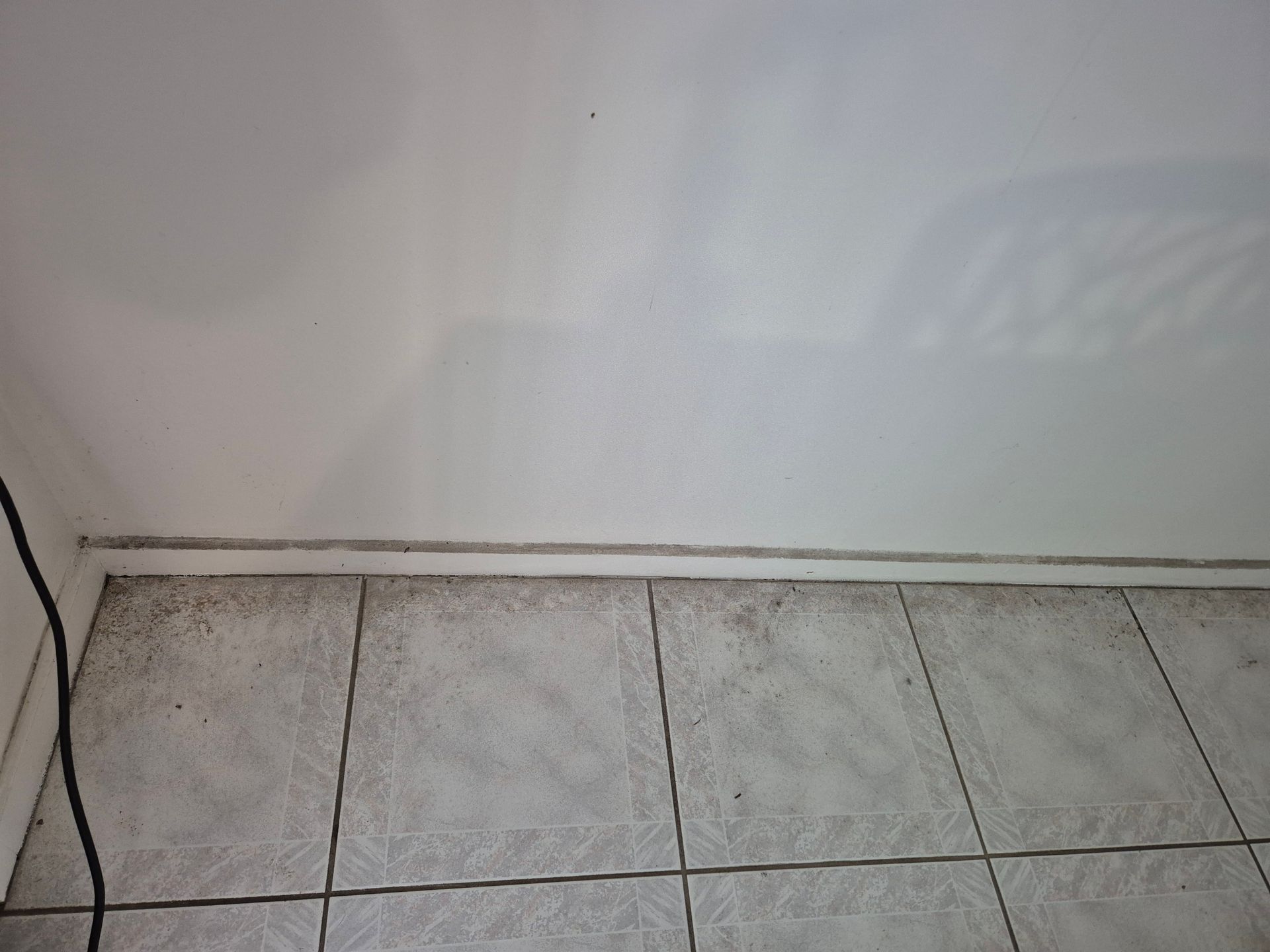 Before: White Wall Meeting Tiled Floor, Dirty Grout and Baseboard. Black Cord on Left — KirraLina Cleaning Services in Banora Point, NSW