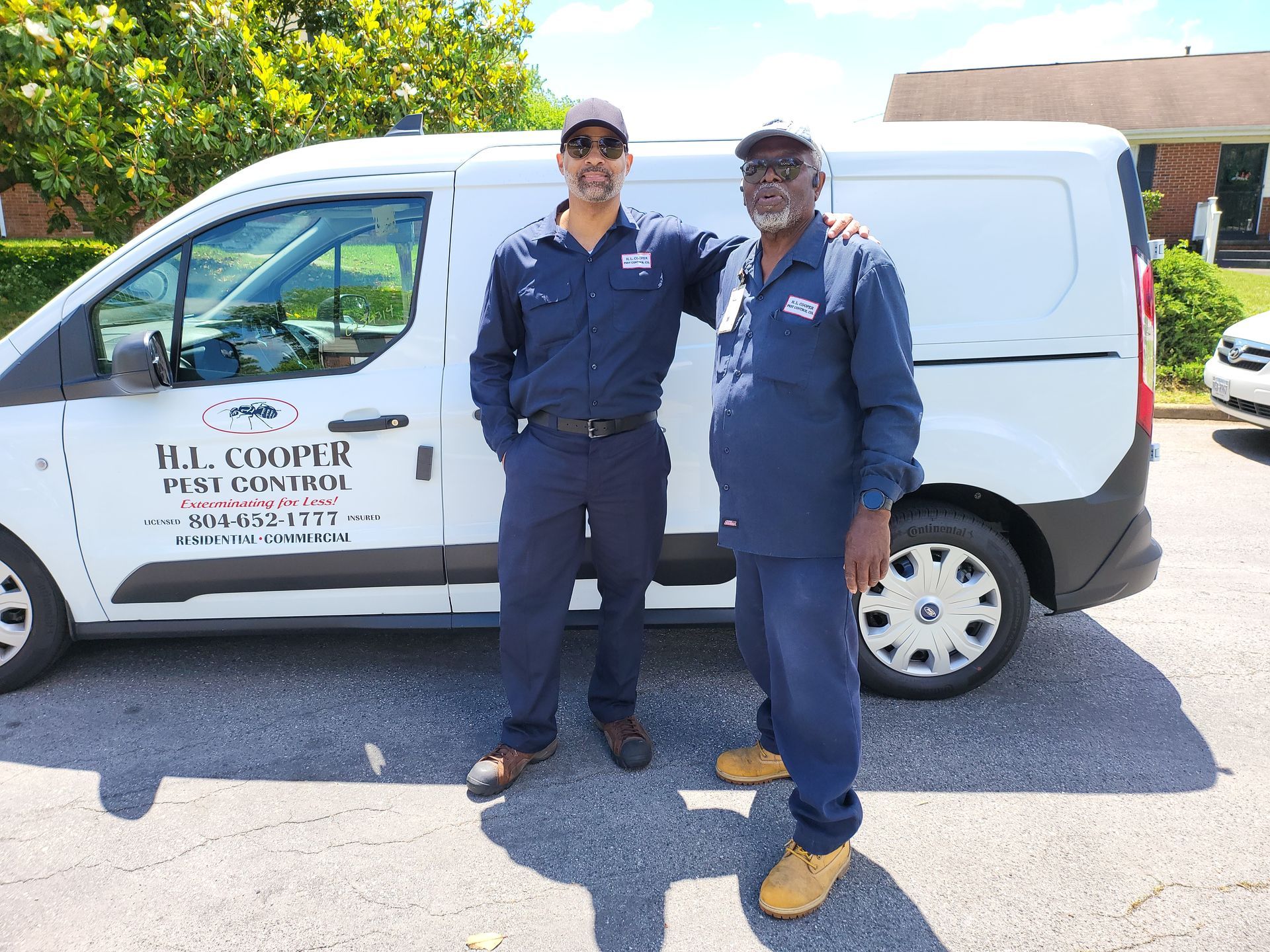 Two men in blue work uniforms stand next to a white van with business logo.