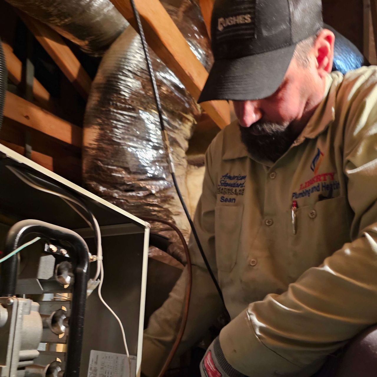 HVAC technician in a crawl space, inspecting a furnace. He's wearing a company uniform and cap.